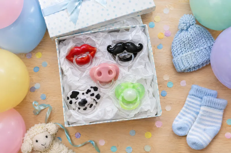 KI-Bild: Geschenkbox mit lustigen Schnullern – Lippen, Schnurrbart, Schweinenase, Kuhflecken – zwischen Luftballons und Babysocken